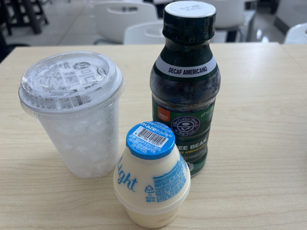 Ingredients to make the viral Ttungba Latte: a packaged plastic ice cup, a pouch of unsweetened Black Americano, and a classic jar-shaped Binggrae Banana Flavored Milk from a Korean convenience store.