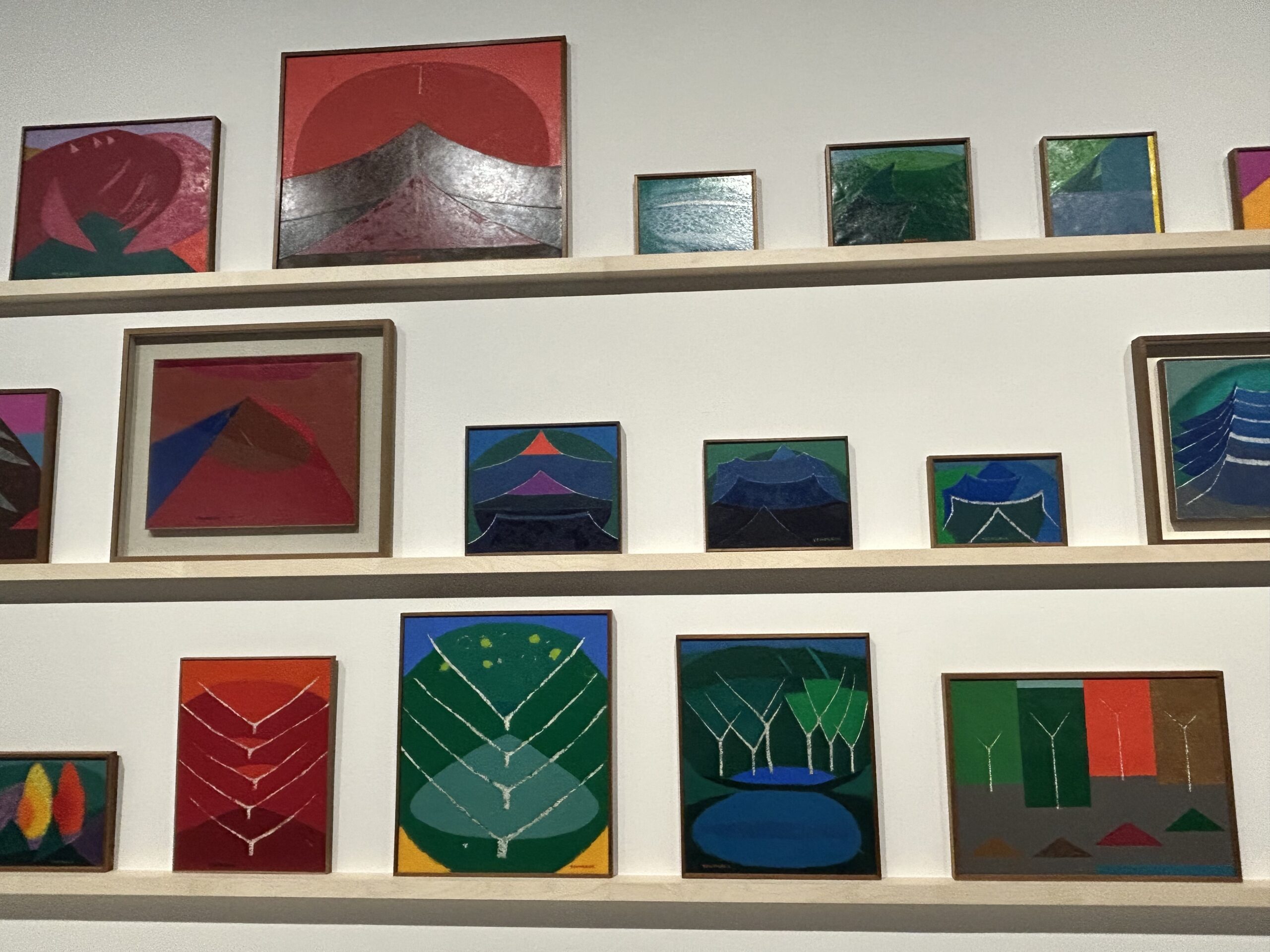A photograph of colorful abstract paintings by Yoo Youngkuk displayed on walls within a white, modern museum exhibition hall. The artworks features his signature bright colors and geometric shapes representing mountains.